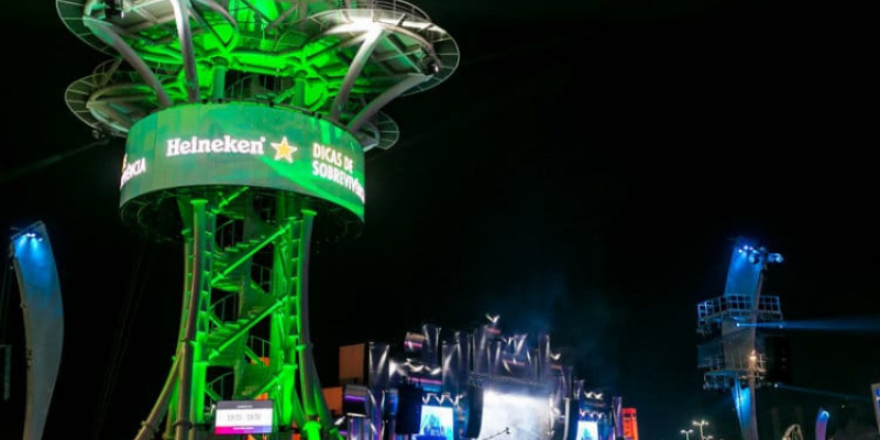 Foto de ativação da Heineken no Rock in Rio 2019, em que se mostra o céu em início de noite, em que uma pessoa participa da tirolesa da marca, com um letreiro em led esverdeado em que se lê “E a diversão”
