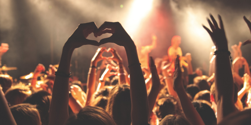 Foto de um show em que o público está com as mãos para cima e algumas pessoas fazem o símbolo de coração.