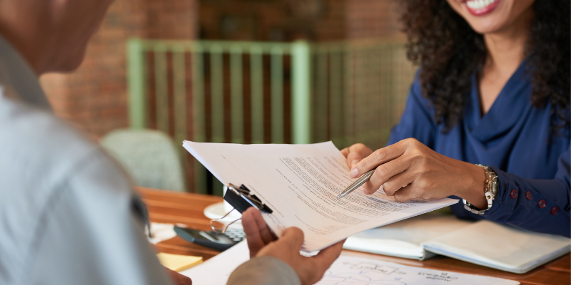 profissional mulher sorridente entregando um documento com caneta para assinatura para um profissional homem em uma mesa com xícara de café